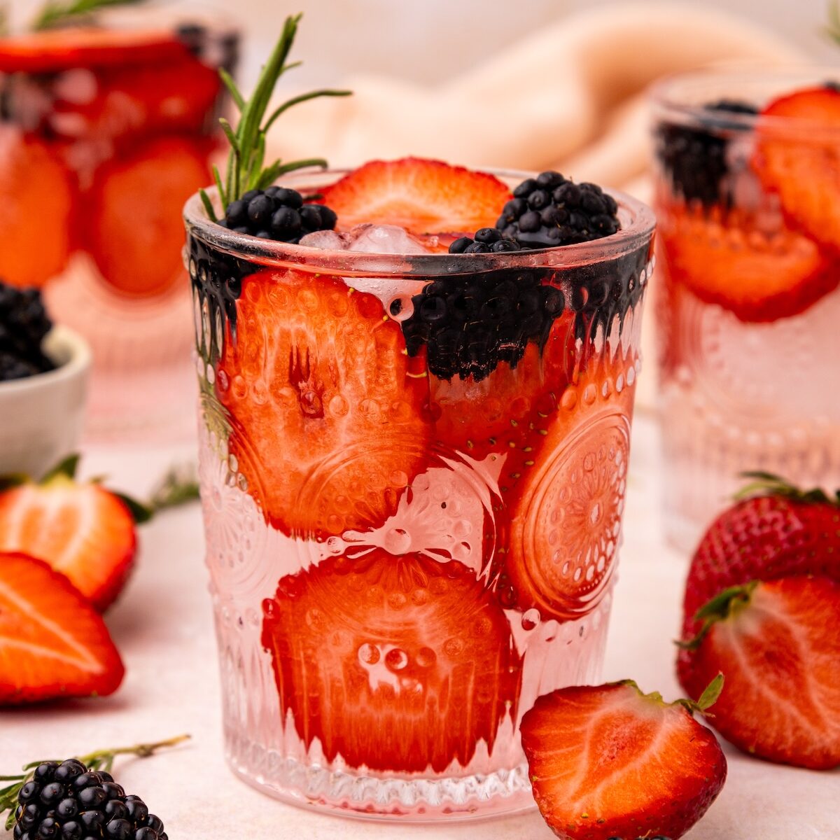 A glass of strawberry water with ice, blackberries, and a fresh rosemary garnish.
