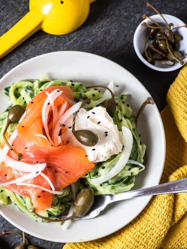 Avocado Cucumber Noodles Topped with Smoked Salmon Ribbons in a white bowl places on a yellow kitchen towel. 
