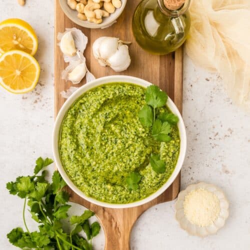 Bowl of cilantro pesto with cashews, garlic, oil, lemon, and cheese on a board.