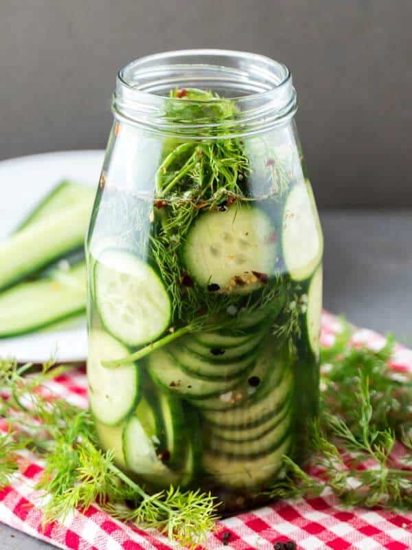 Side shot of a tall glass jar filled with cucumber slices, fresh herbs and spices. Sitting on top a red checkered kitchen towel. Fresh herbs surrounds it.