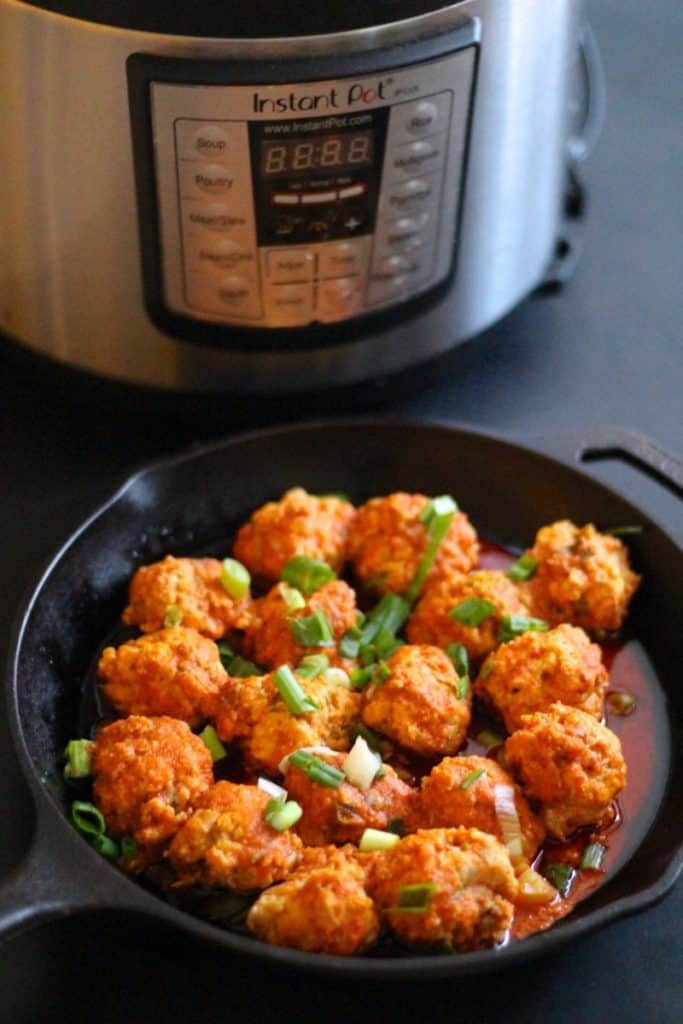 Keto instant pot buffalo chicken meatballs topped with fresh spring onion in a skillet next to an instant pot on a black surface.