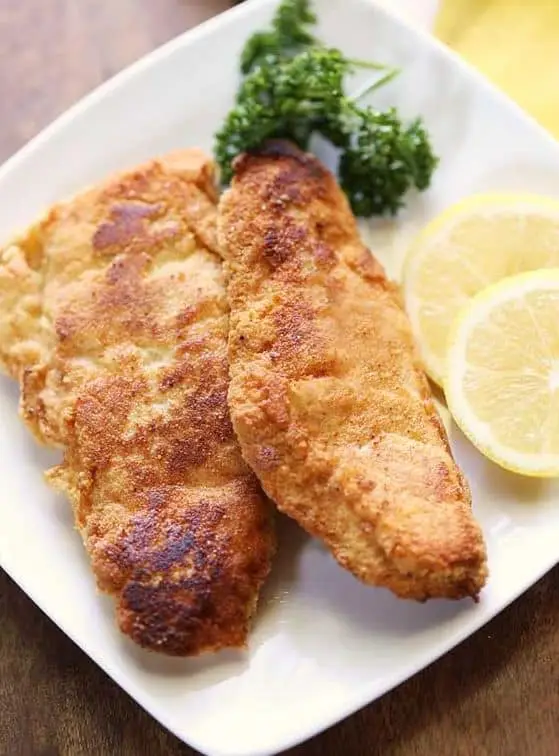 Keto fried fish with fresh lemon slices and parsley in a white plate with a wooden background. 