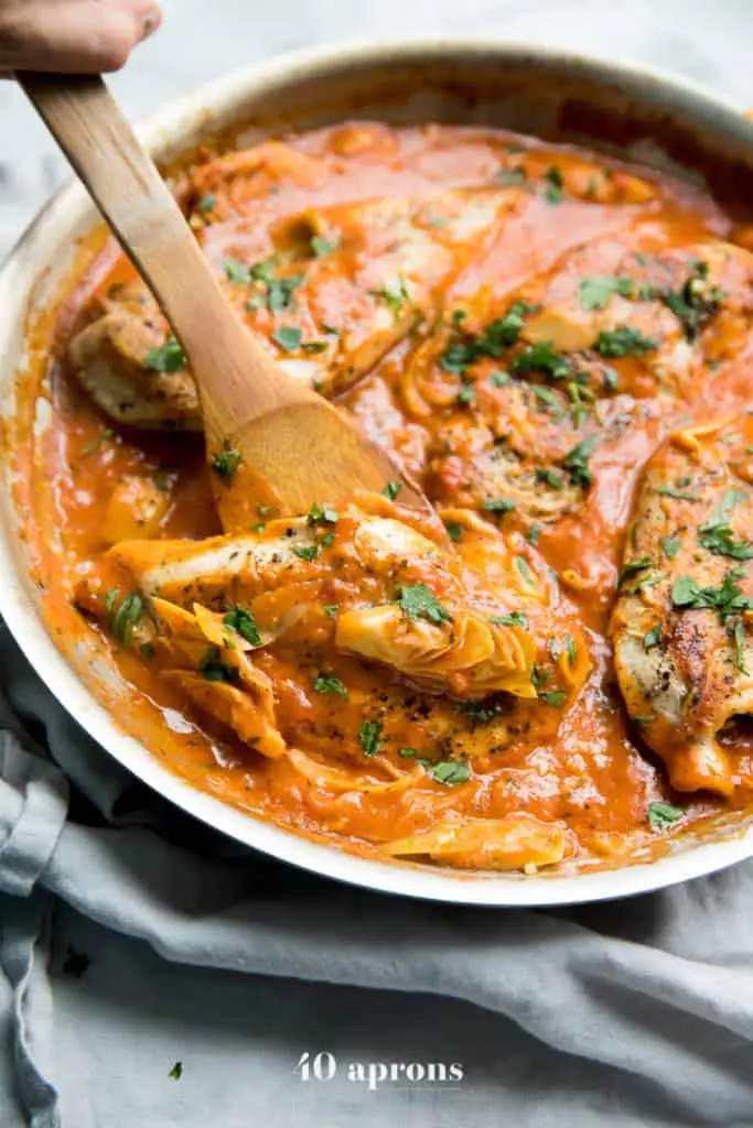 Chicken and artichokes in a creamy red tomato sauce topped with chopped fresh herbs in a white bowl with a wooden spoon.