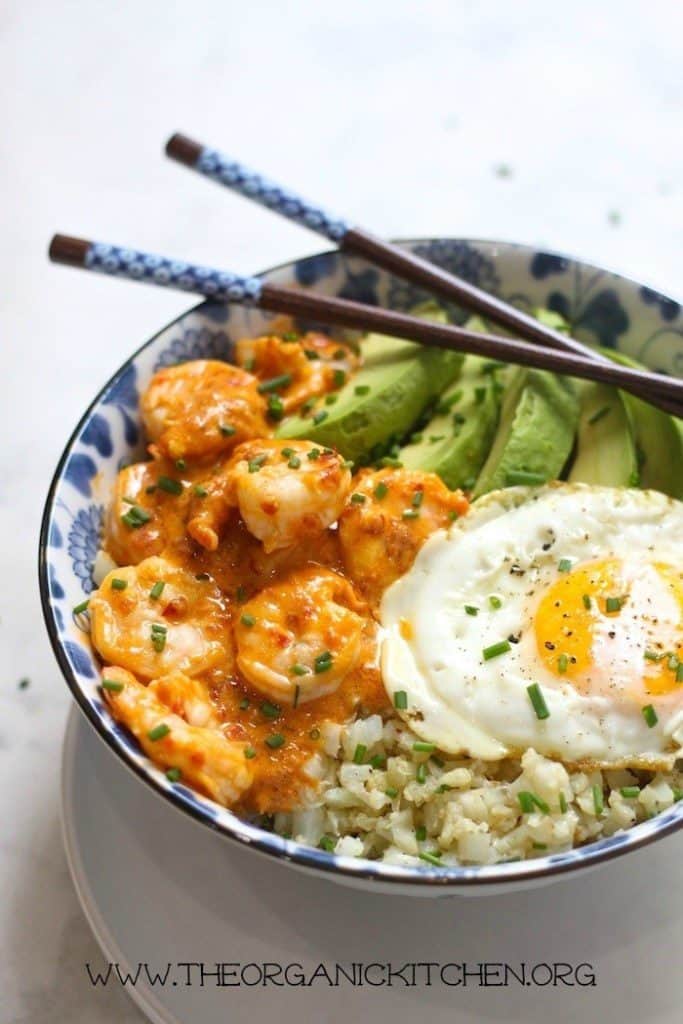 Fried egg, shrimp in creamy sauce, cauliflower rice and sliced avocado in a white and blue bowl with chopsticks on a white background.