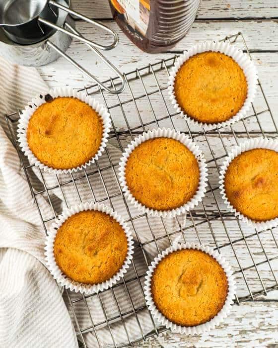 Six coconut flour muffins are arranged on a wire rack placed on a white wooden surface.