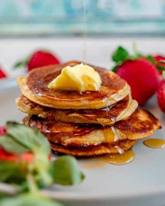 maple syrup being drizzled on coconut flour pancakes. strawberries in the background