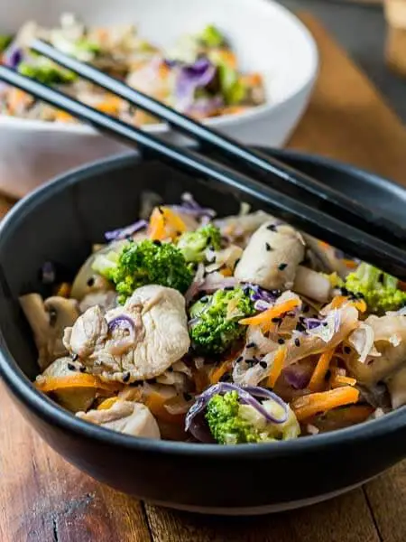 Low top shot of black bowl filled with stir fried chicken and vegetables and black chopsticks.