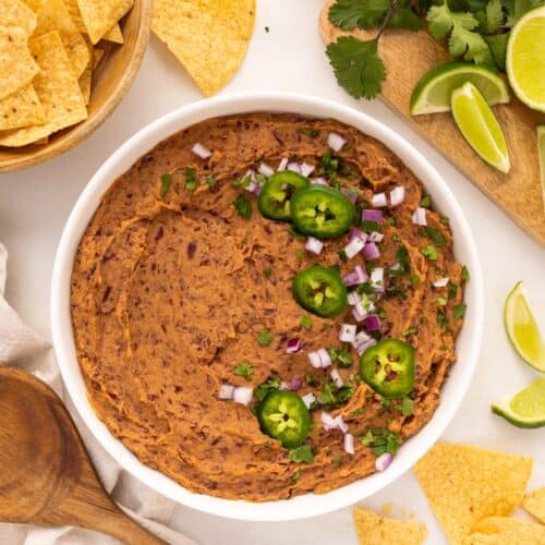 A bowl of refried kidney beans topped with jalapeños, onions, cilantro, chips, and lime.