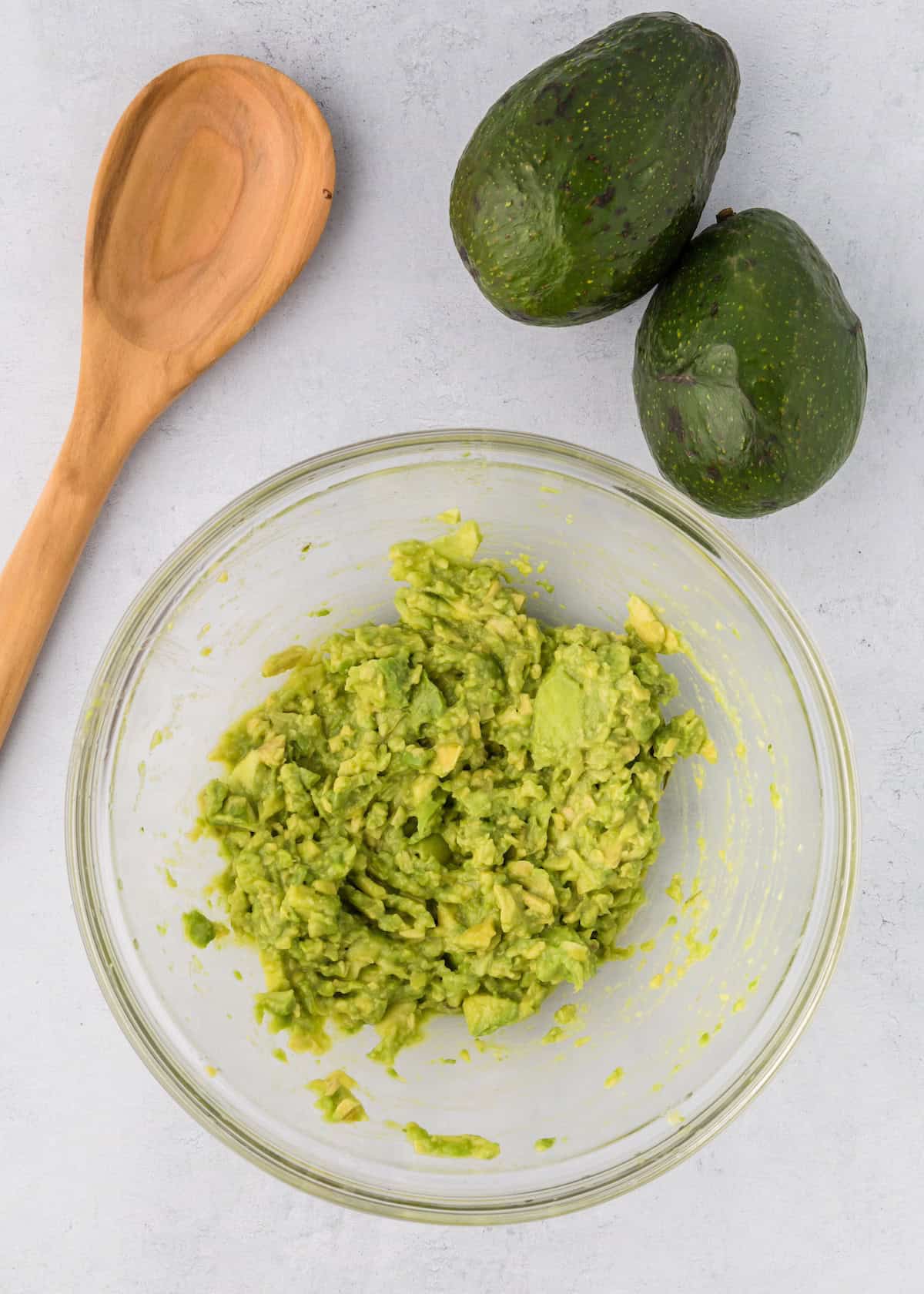 Mashed avocado in a glass bowl, two whole avocados, and Avocado Chicken Salad on a white surface.
