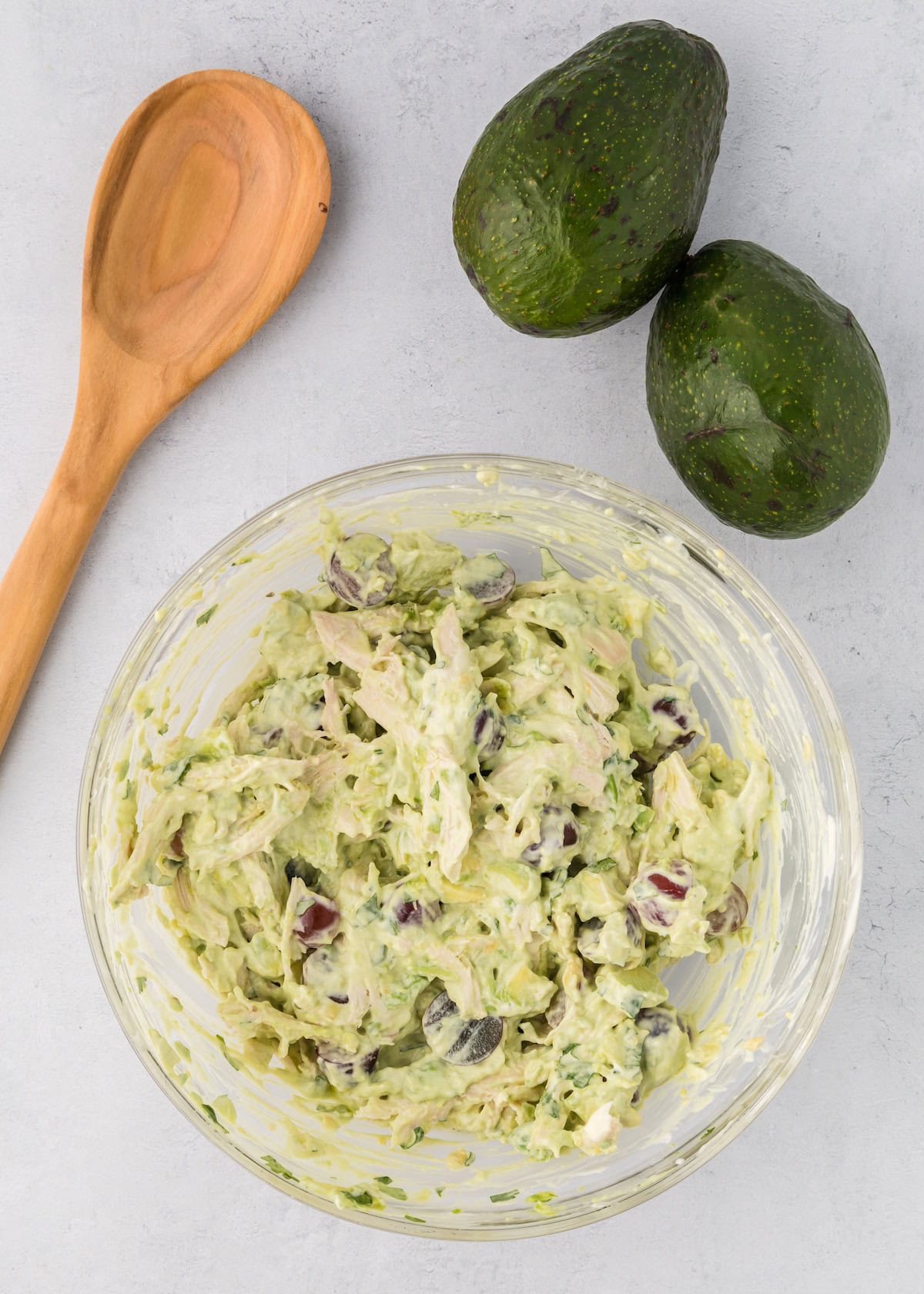 A bowl of Avocado Chicken Salad with grapes, beside whole avocados and a wooden spoon.