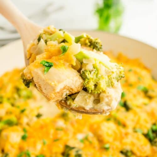 A wooden spoon holds a cheesy chicken broccoli and rice casserole above a casserole dish.