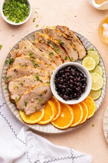 Sliced Cuban pork with orange slices, black beans, lime, and chopped herbs on a plate.