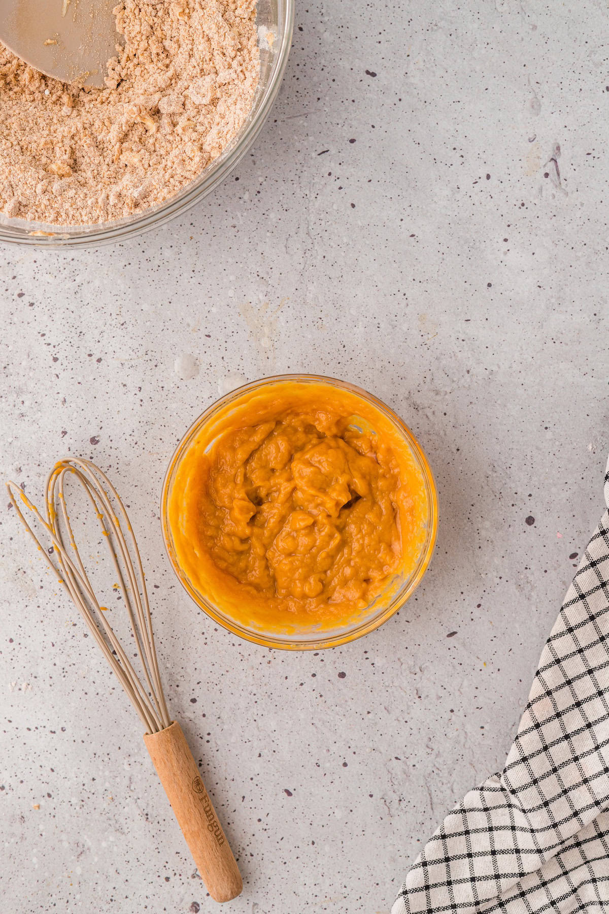 Bowl of orange batter for Gluten-Free Pumpkin Scones, whisk, and flour on a light countertop.