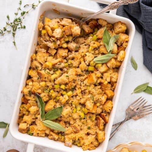 A casserole dish of sourdough stuffing, garnished with fresh sage and served with utensils.