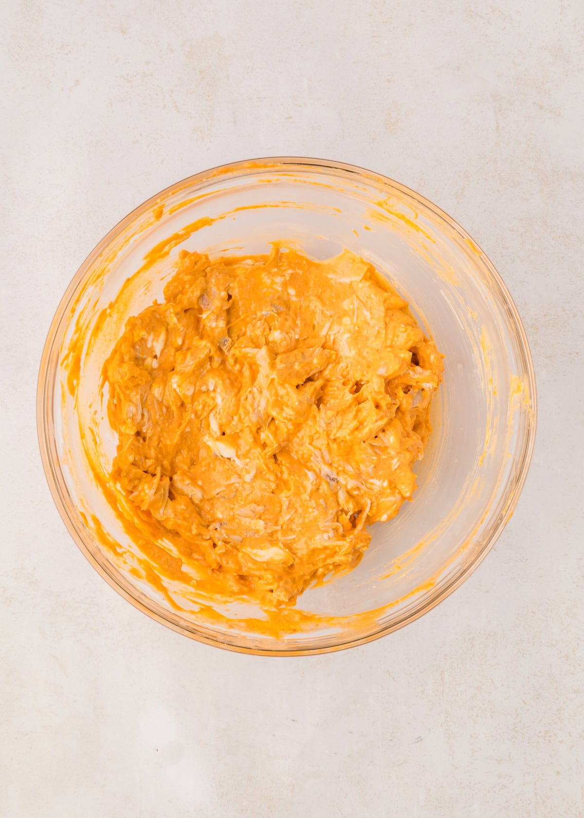 A glass bowl filled with orange, creamy Buffalo Chicken Stuffed Peppers mixture on a light surface.