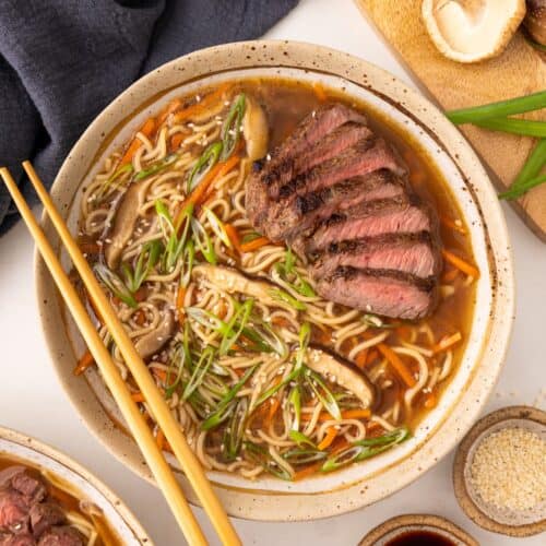 Bowl of beef ramen with sliced steak, mushrooms, green onions, and chopsticks on the side.