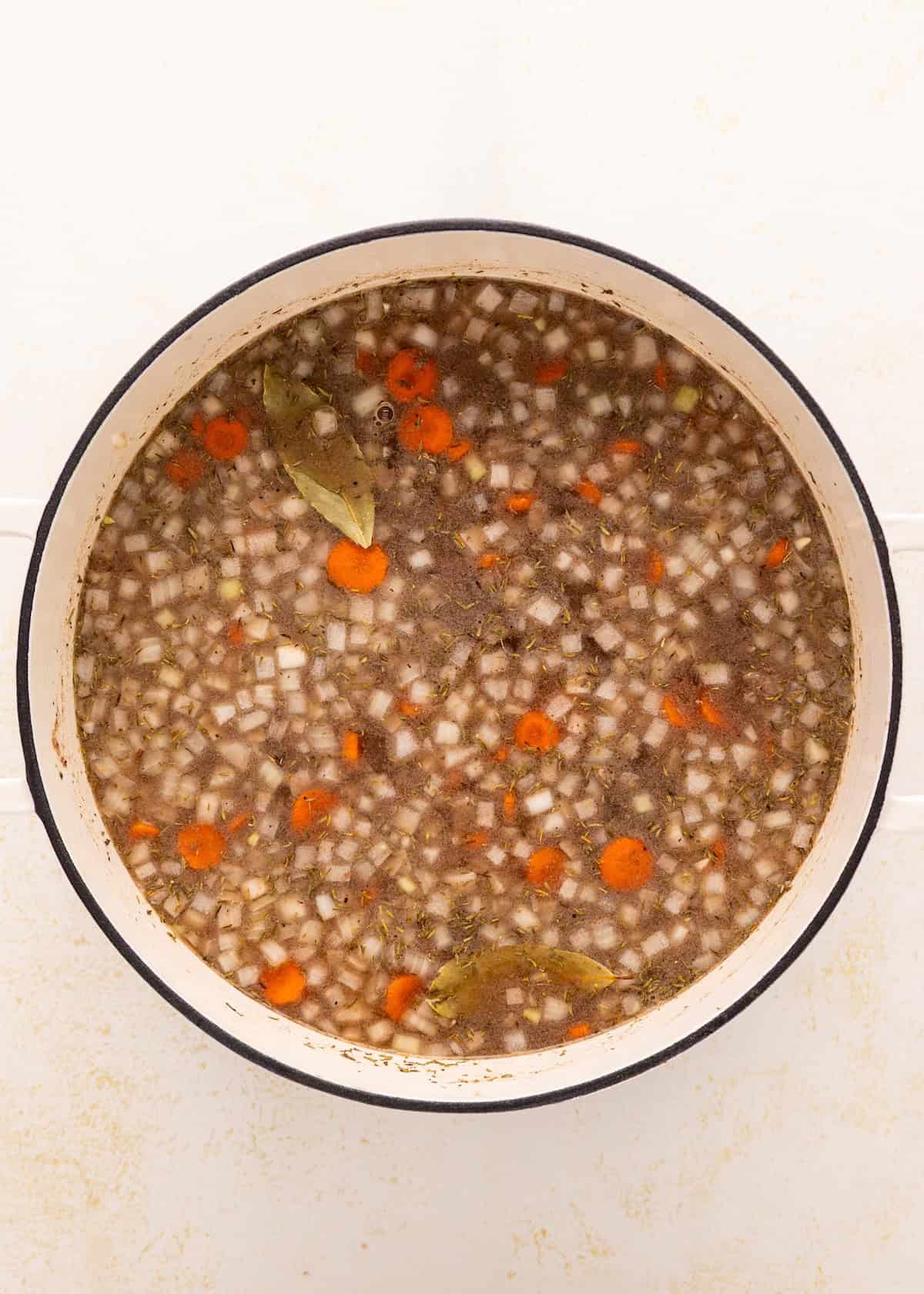 A pot of beef vegetable stew with carrots, onions, herbs, and bay leaves on a white surface.
