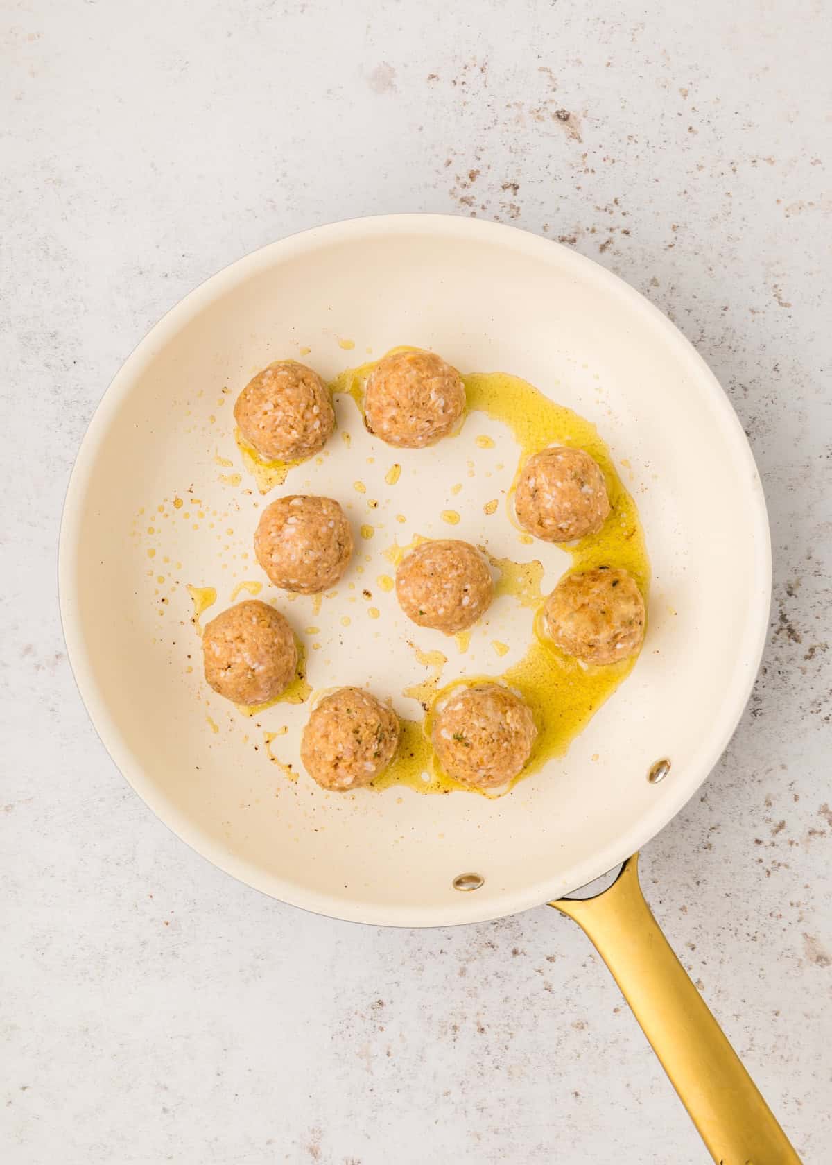 Nine uncooked BBQ Chicken Meatballs in a white frying pan with oil on a light countertop.