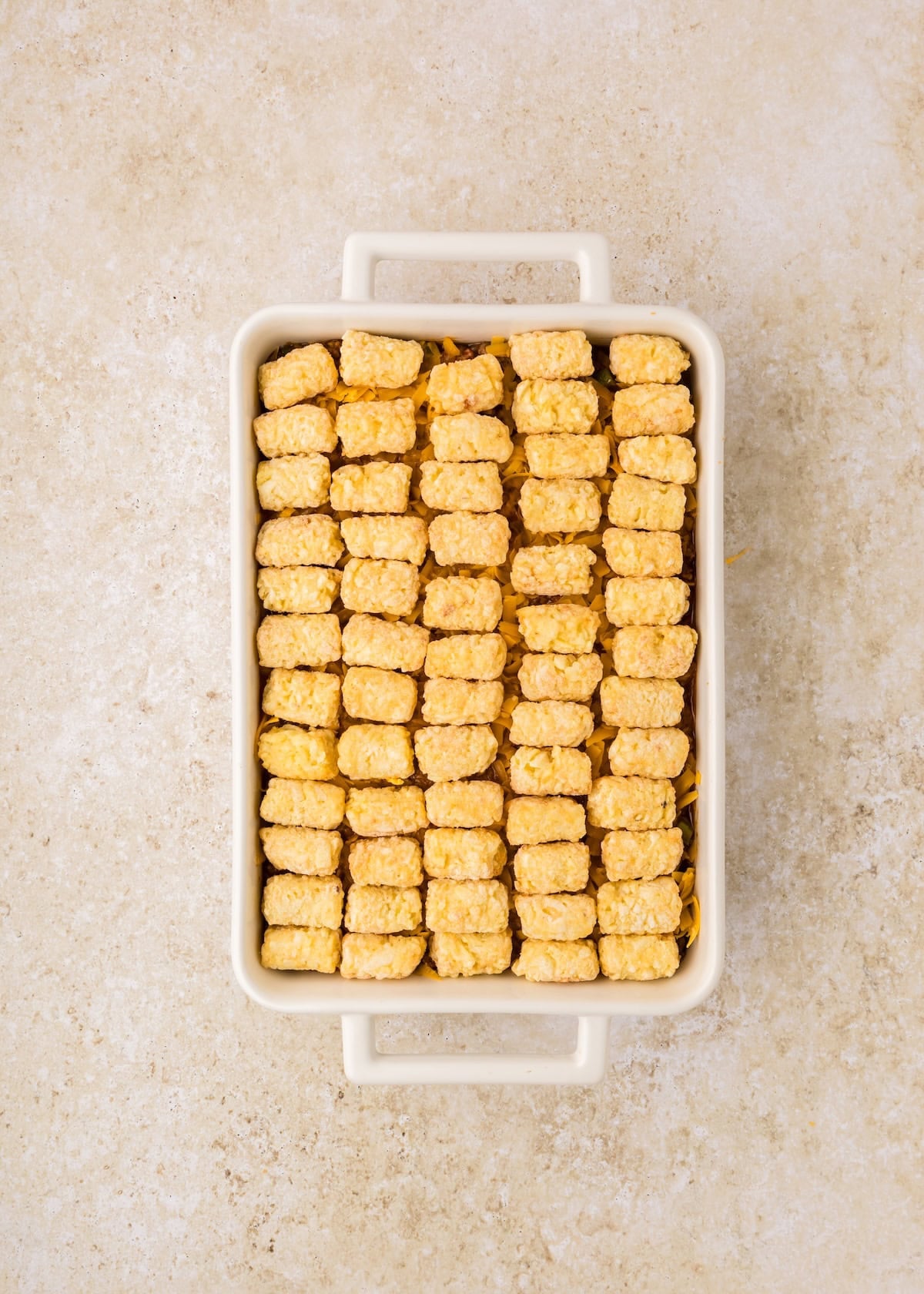 A baking dish of Sloppy Joe Casserole topped with rows of frozen tater tots on a beige surface.