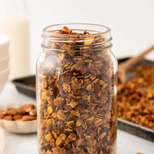 A glass jar of vanilla almond granola sits on a white surface near a baking tray.