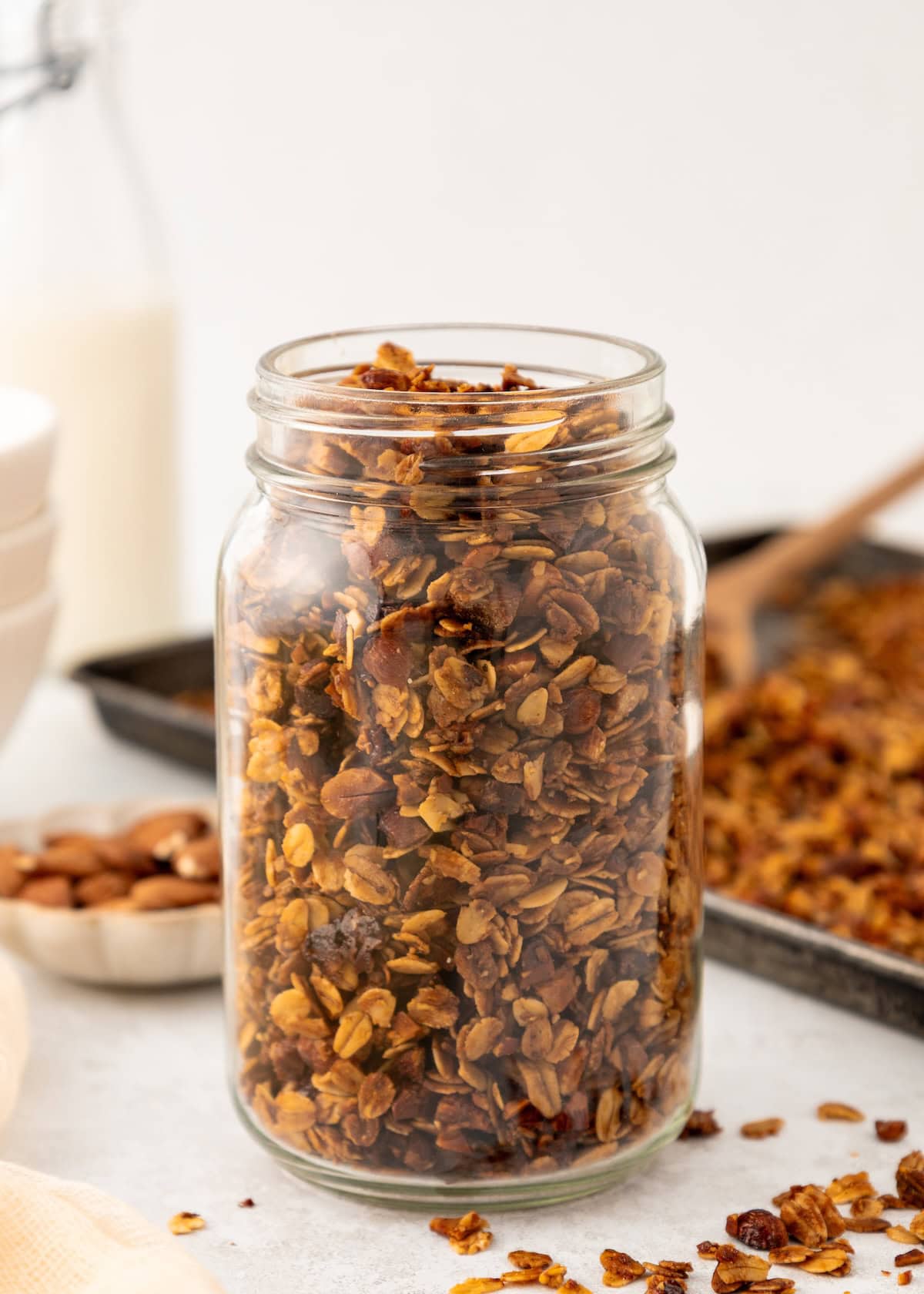 A glass jar of vanilla almond granola sits on a white surface near a baking tray.