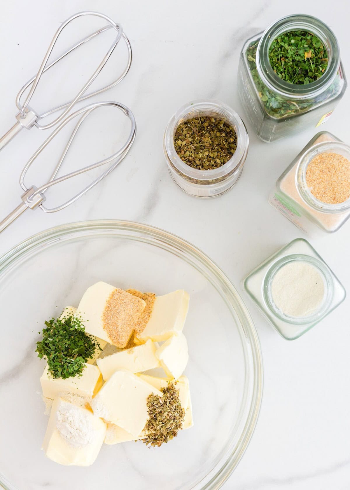 Butter, herbs, and seasonings in a bowl with a hand mixer and spice jars on a white surface.