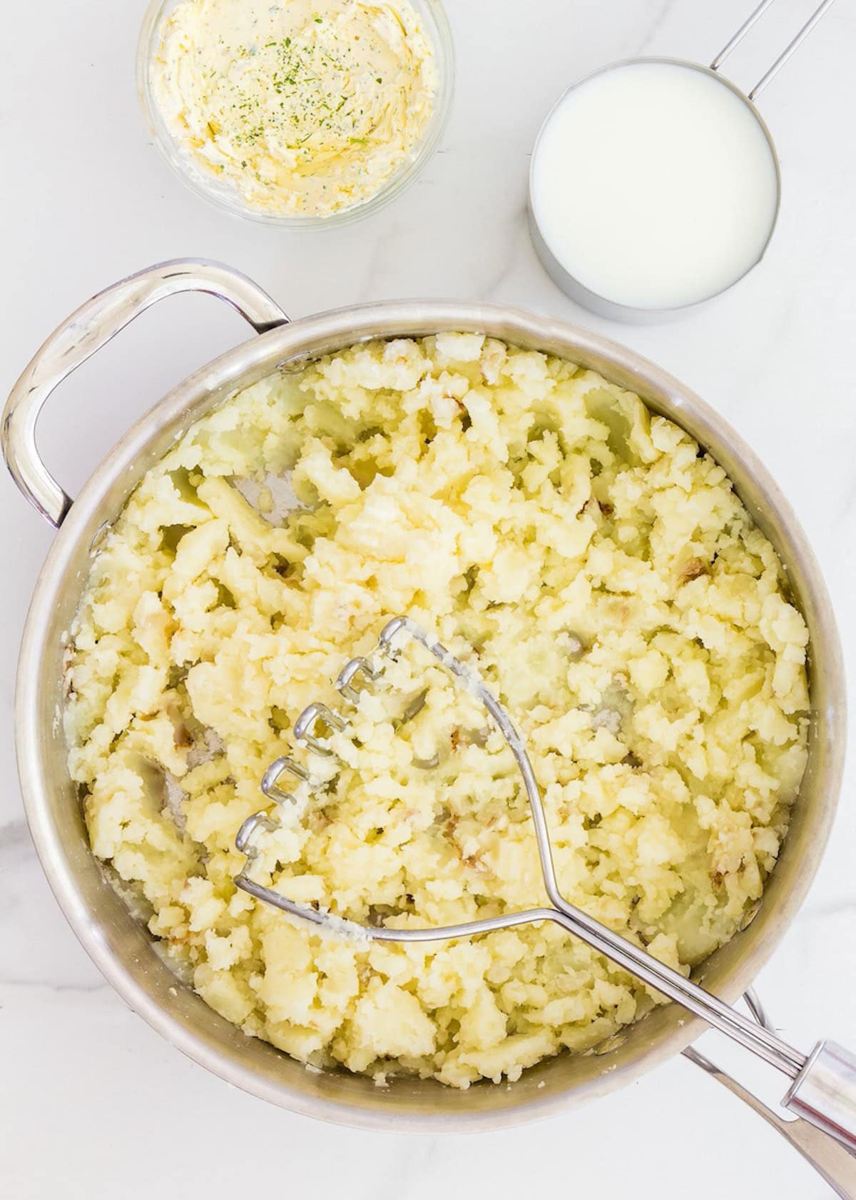 Mashed potatoes in a pot with a masher, beside a cup of milk and a bowl of seasoned butter.