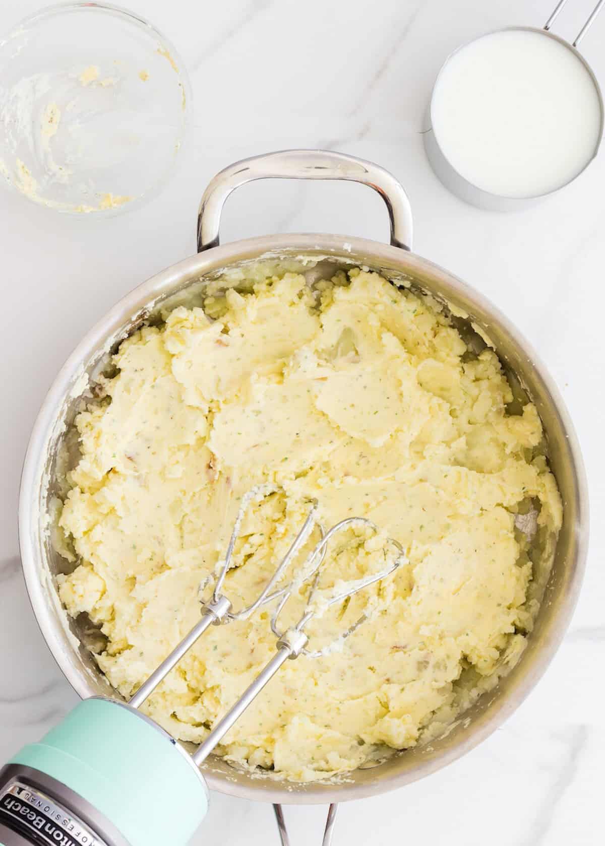A bowl of mashed potatoes with an electric mixer and a cup of milk nearby.