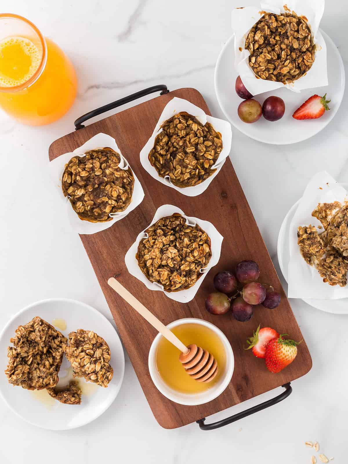 Oat muffins on a wooden tray with honey, grapes, strawberries, and a glass of orange juice.