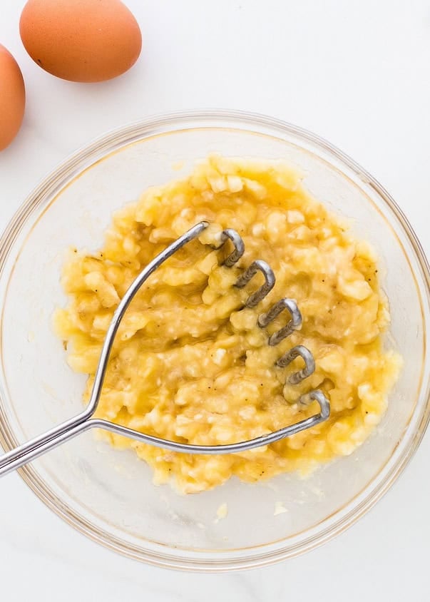 Mashed banana in a glass bowl with a metal masher, eggs nearby on a white surface.