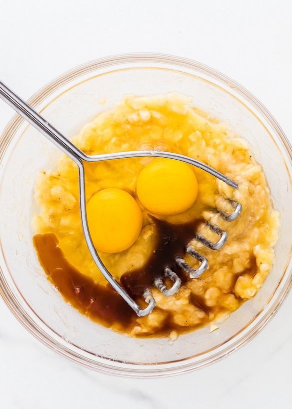 A bowl with eggs, mashed bananas, vanilla, and a masher on a light surface.
