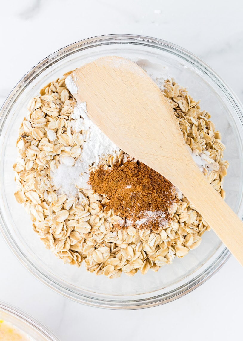 A glass bowl with oats, cinnamon, baking soda, and a wooden spoon on top.