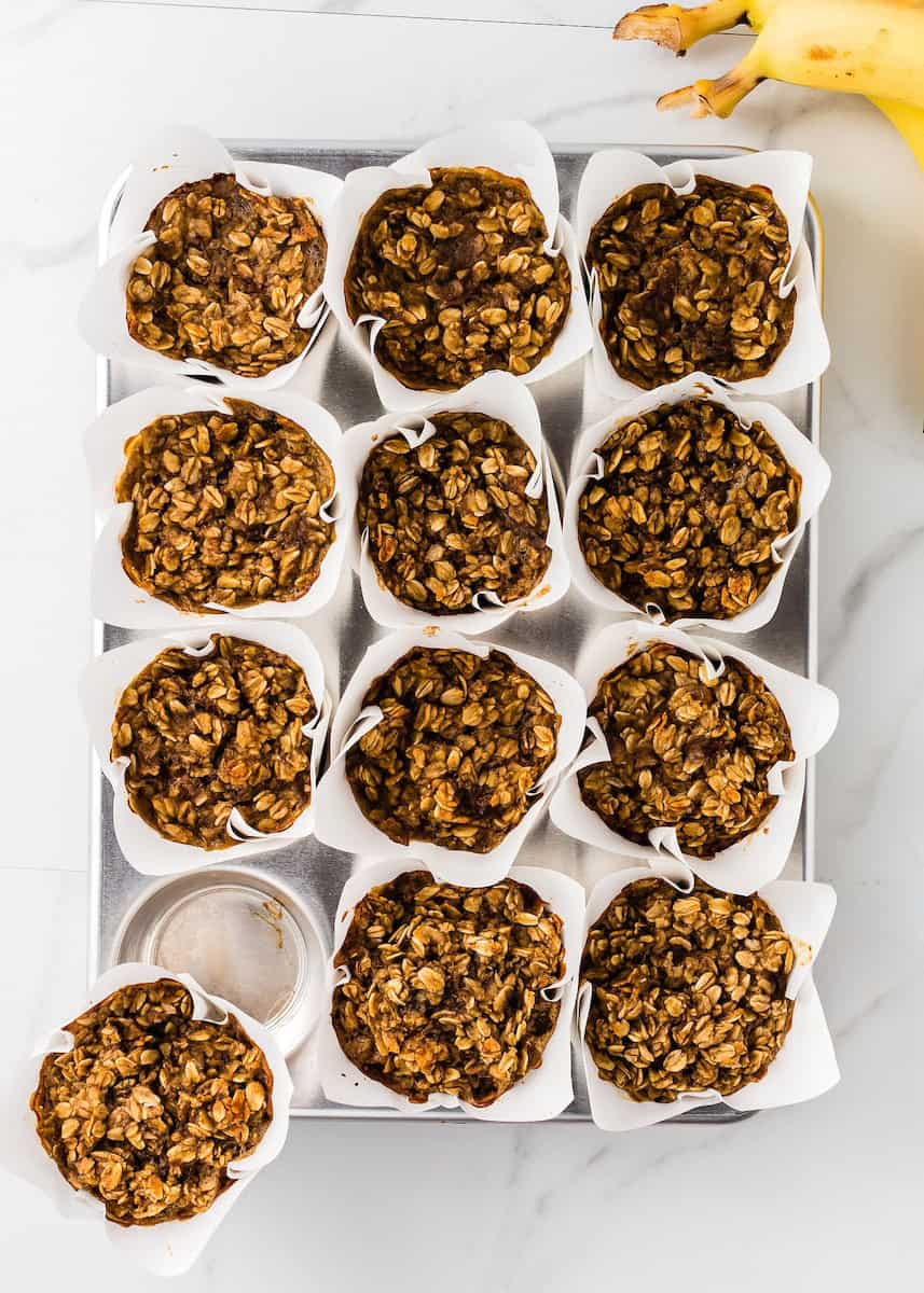 A tray of twelve oatmeal muffins in paper liners, with bananas visible in the corner.