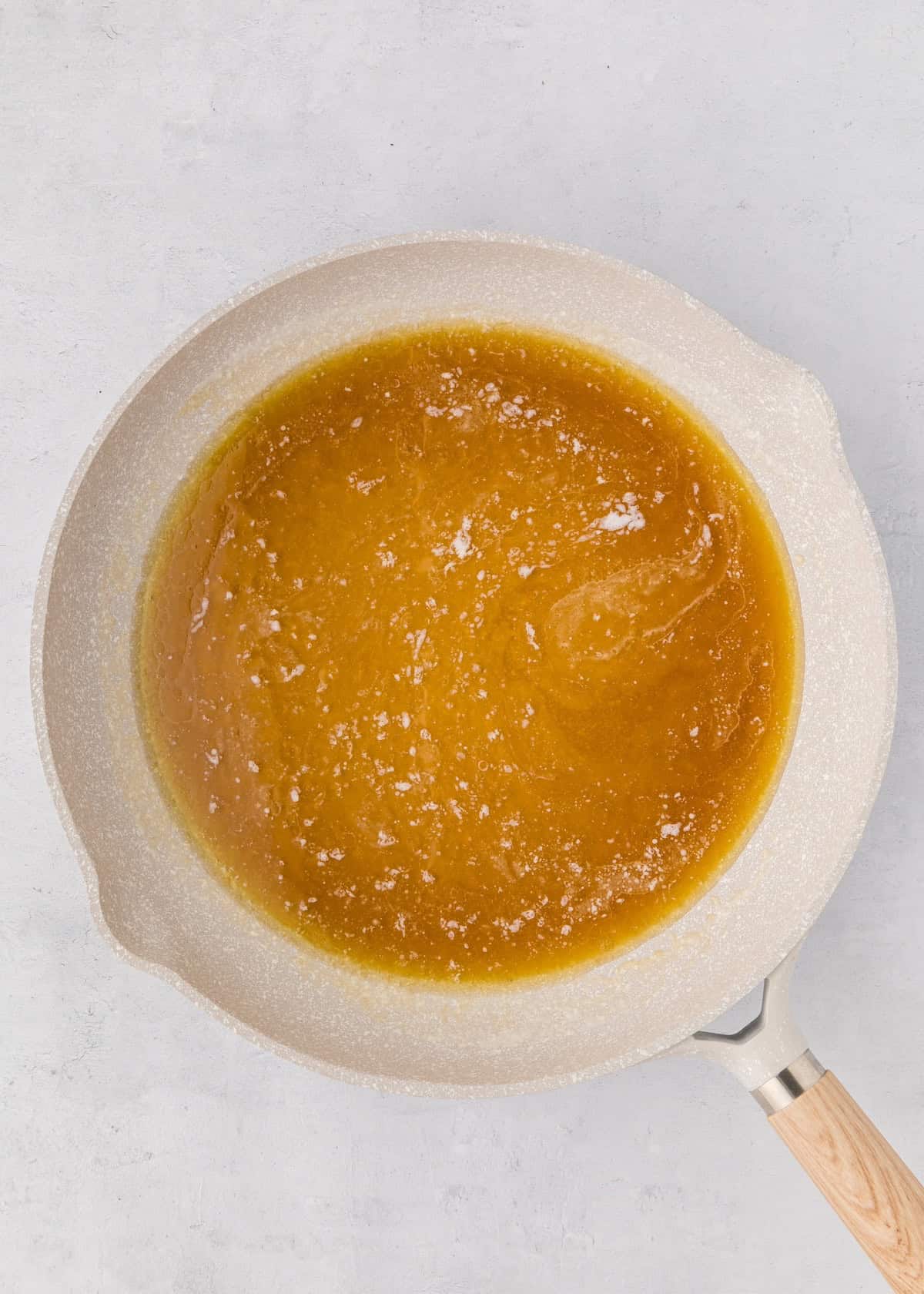 Light brown melted sugar in a white frying pan with a wooden handle, on a gray background.