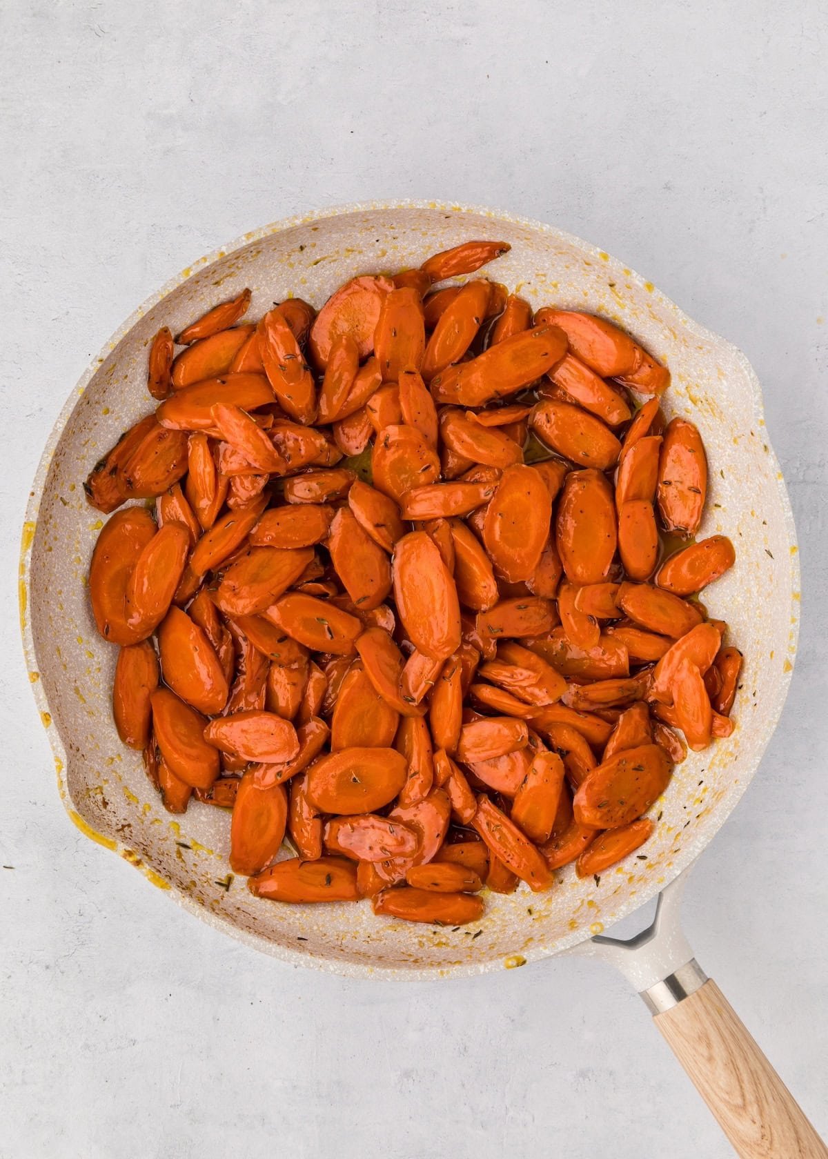 Sliced cooked carrots in a skillet with a light wooden handle on a white surface.