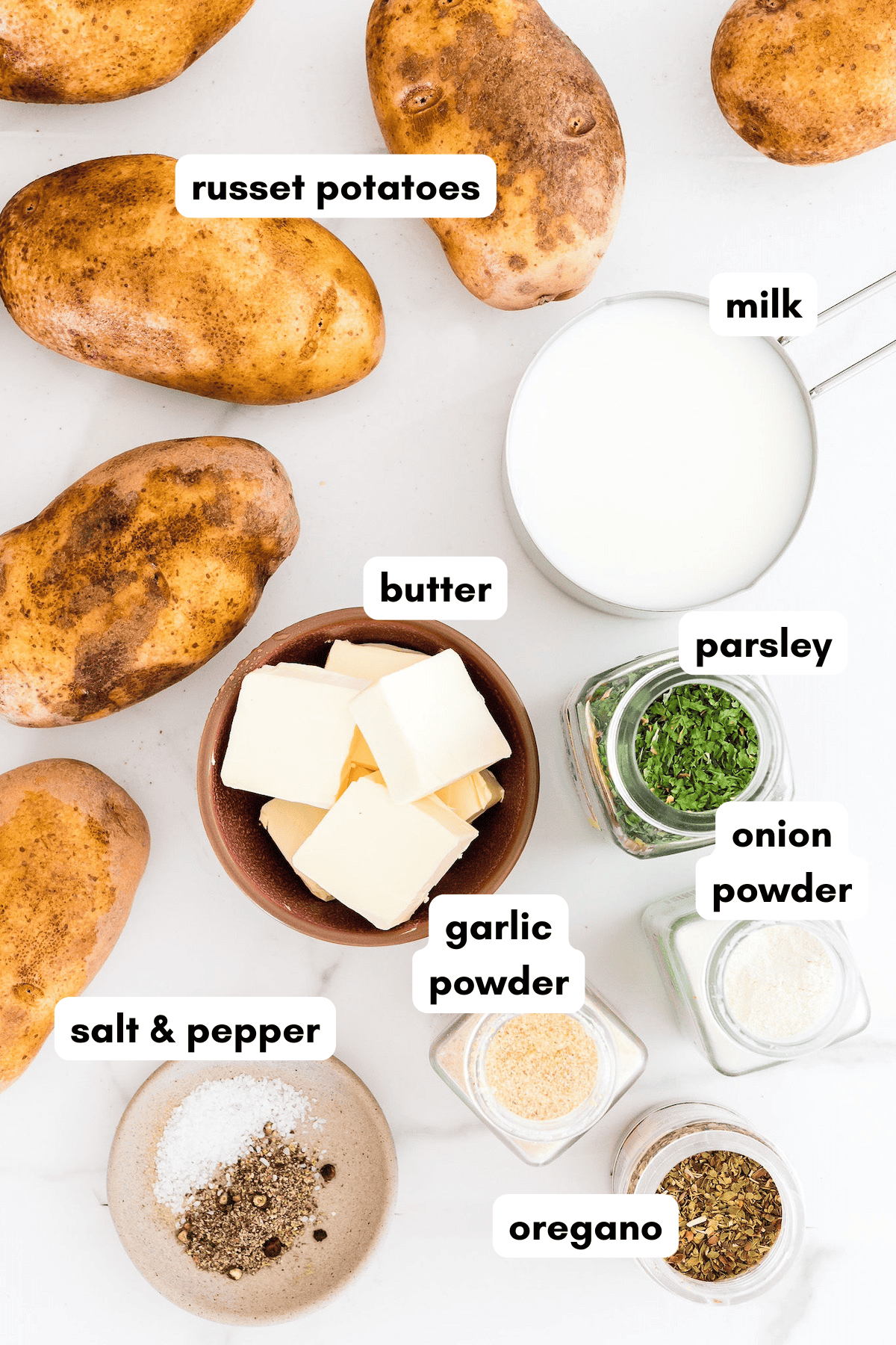 Overhead view of russet potatoes, milk, butter, spices, and seasonings on a white surface.