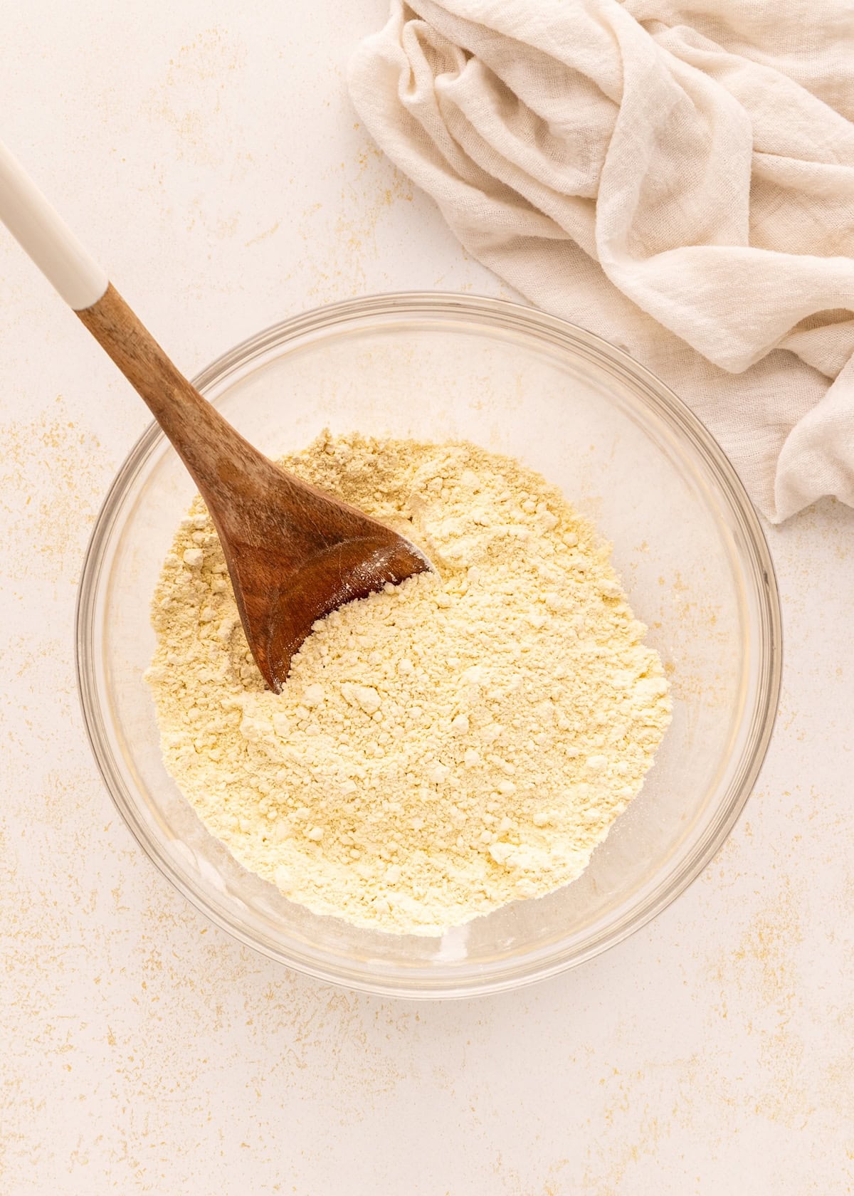 A glass bowl of Greek Yogurt Bagels flour mixture with a wooden spoon on a light surface.