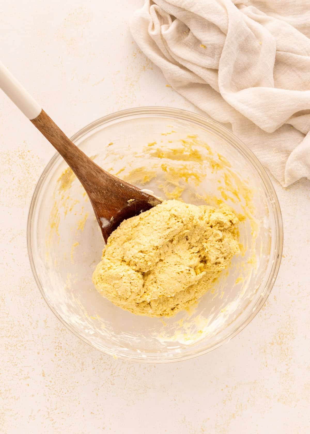 A glass bowl with yellow dough for Greek Yogurt Bagels and a wooden spoon on a light surface.