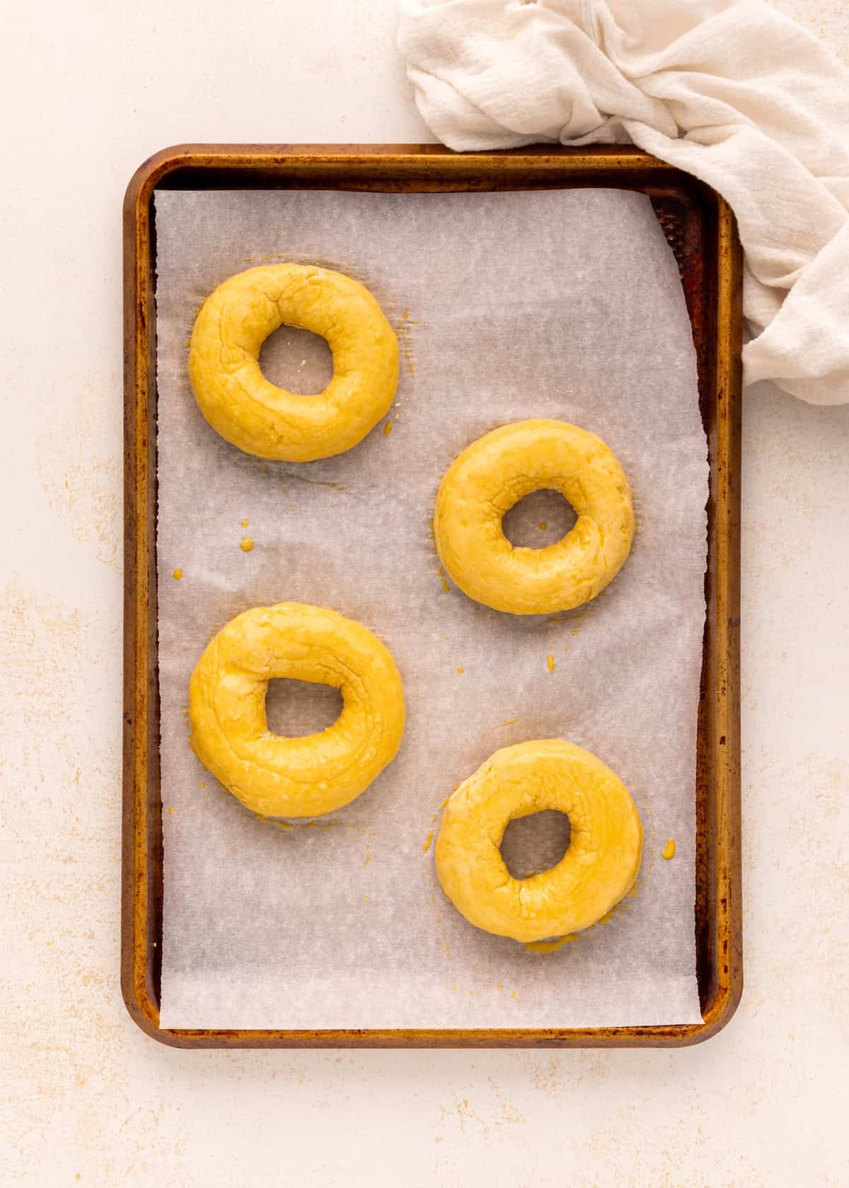 Four unbaked Greek Yogurt Bagels on a parchment-lined sheet, with a white cloth in the corner.
