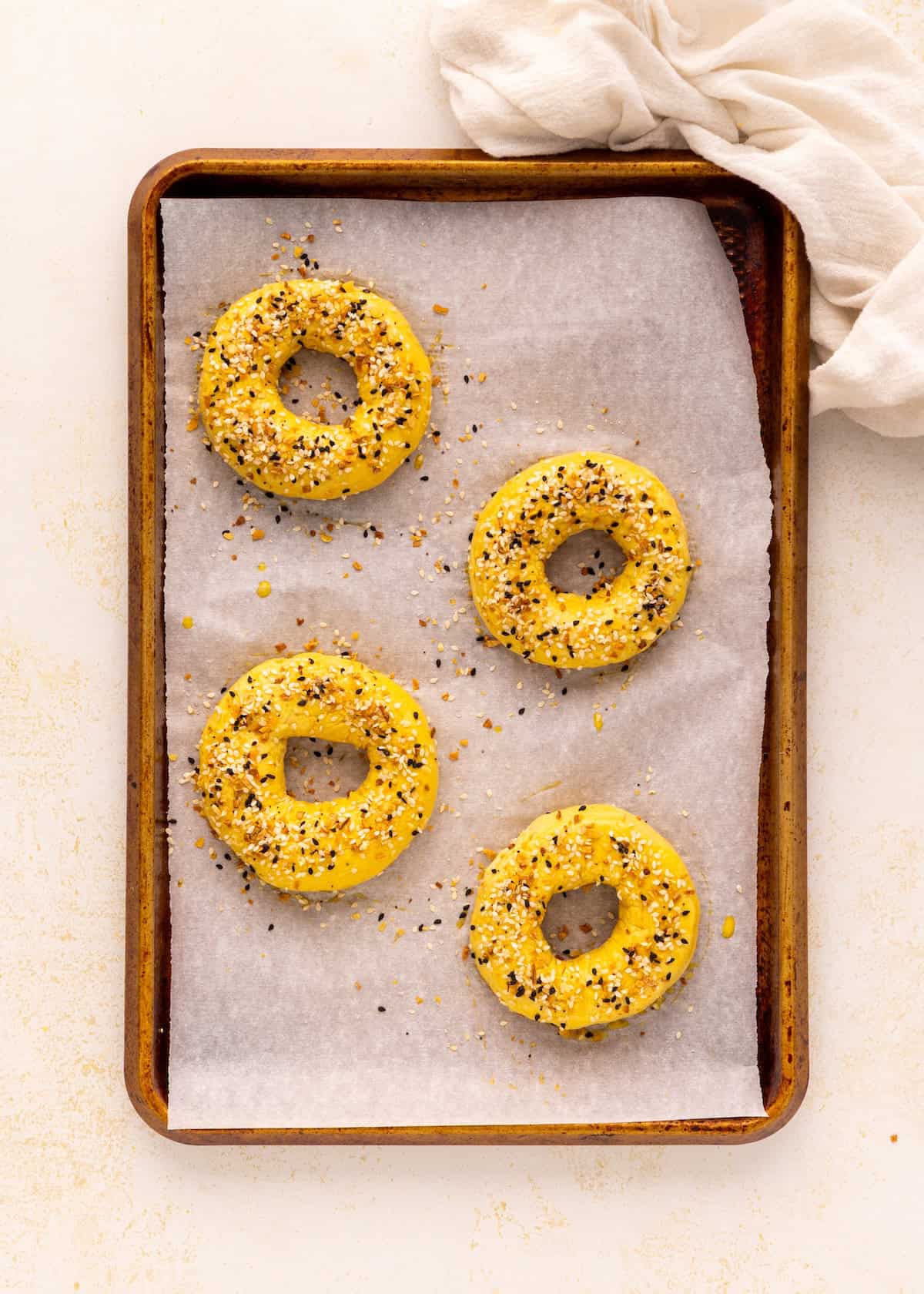 Four unbaked Greek Yogurt Bagels with seasoning on a parchment-lined baking sheet, top view.