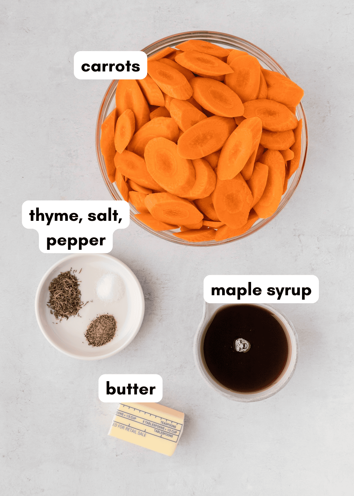 Sliced carrots, maple syrup, butter, thyme, salt, and pepper arranged in bowls on a counter.