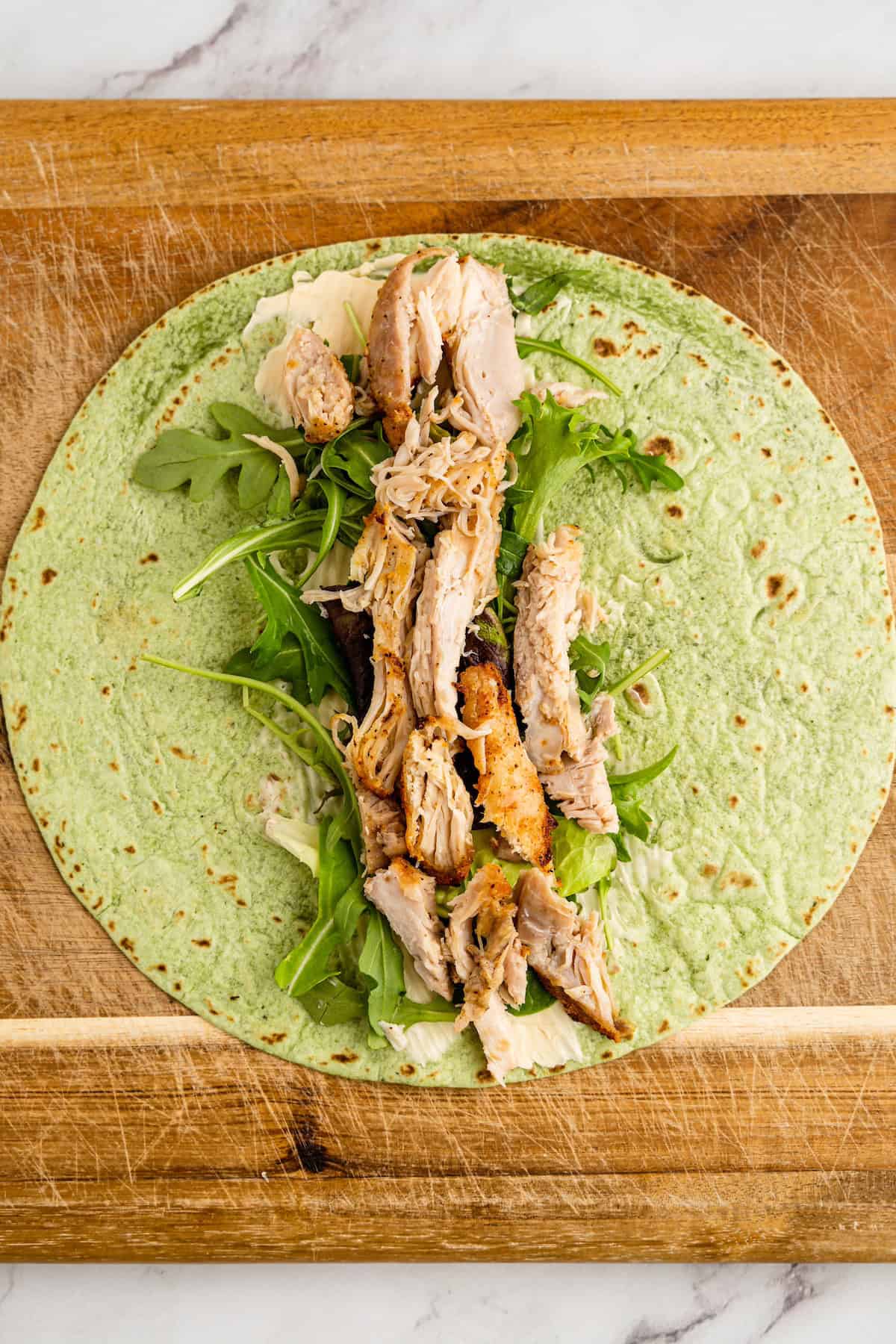 A spinach wrap with arugula, shredded chicken, and cheese on a wooden cutting board.
