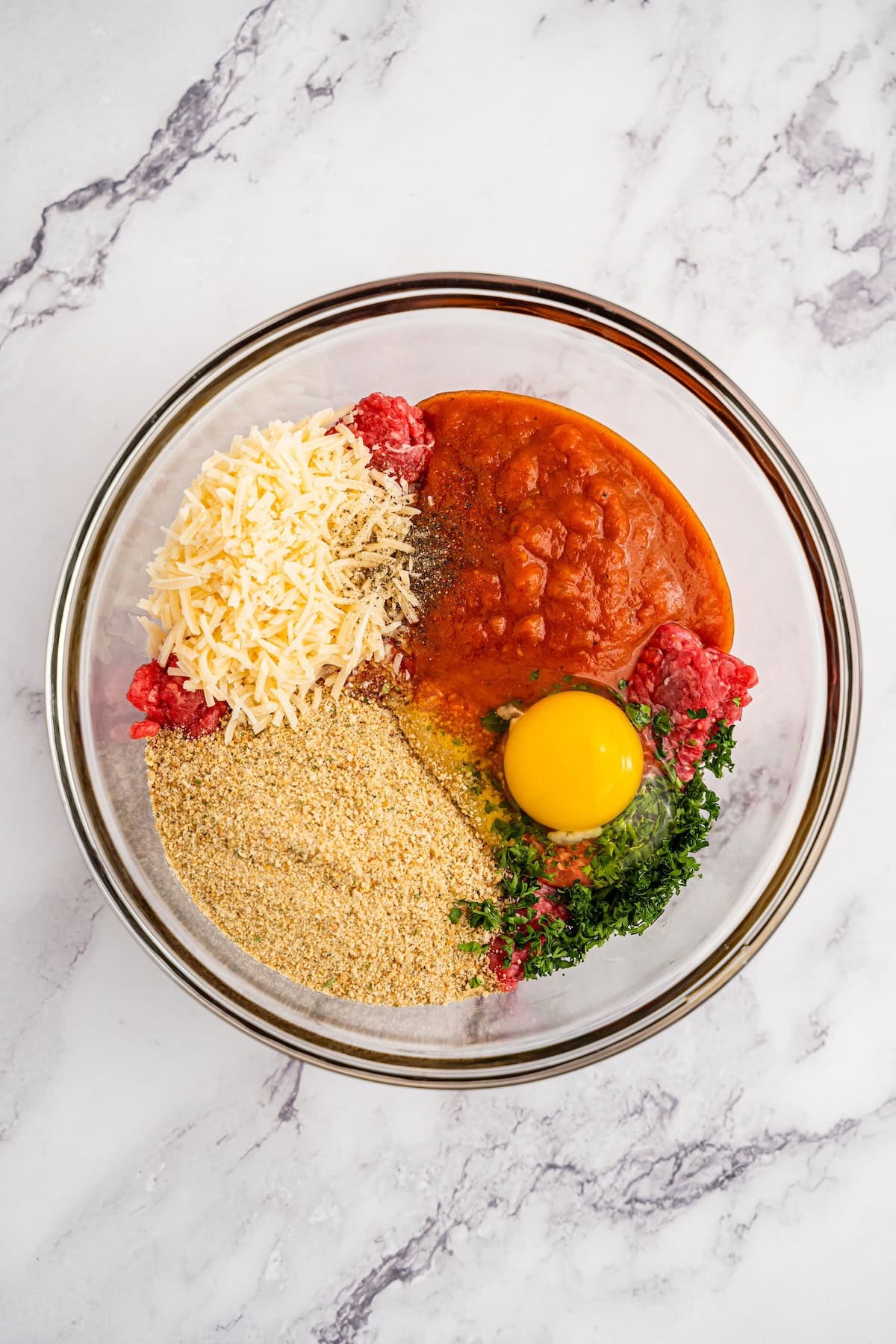 Glass bowl with ground meat, tomato sauce, grated cheese, breadcrumbs, egg, and herbs.