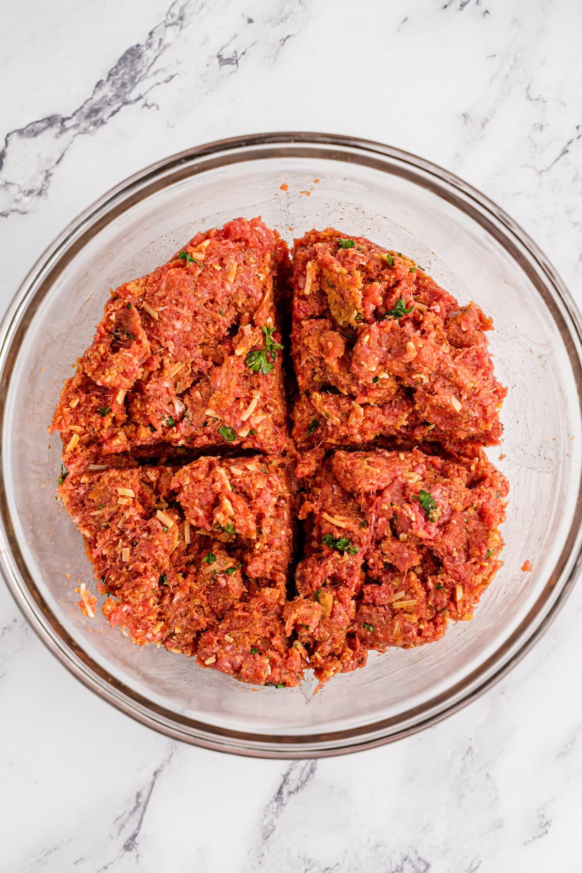Ground meat mixture divided into four sections in a glass bowl on a marble surface.
