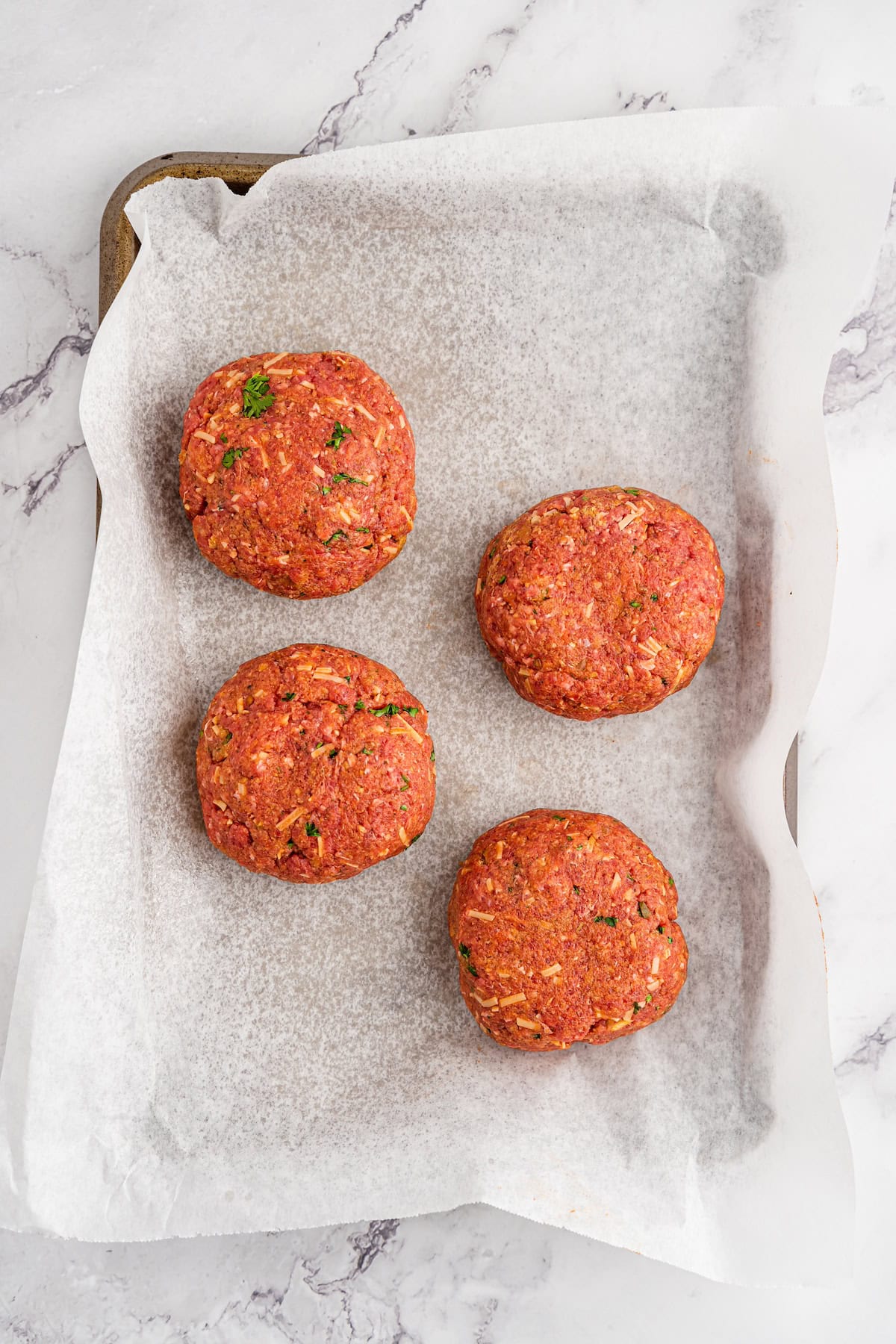 Four raw meat patties with herbs on parchment-lined baking tray, overhead view.