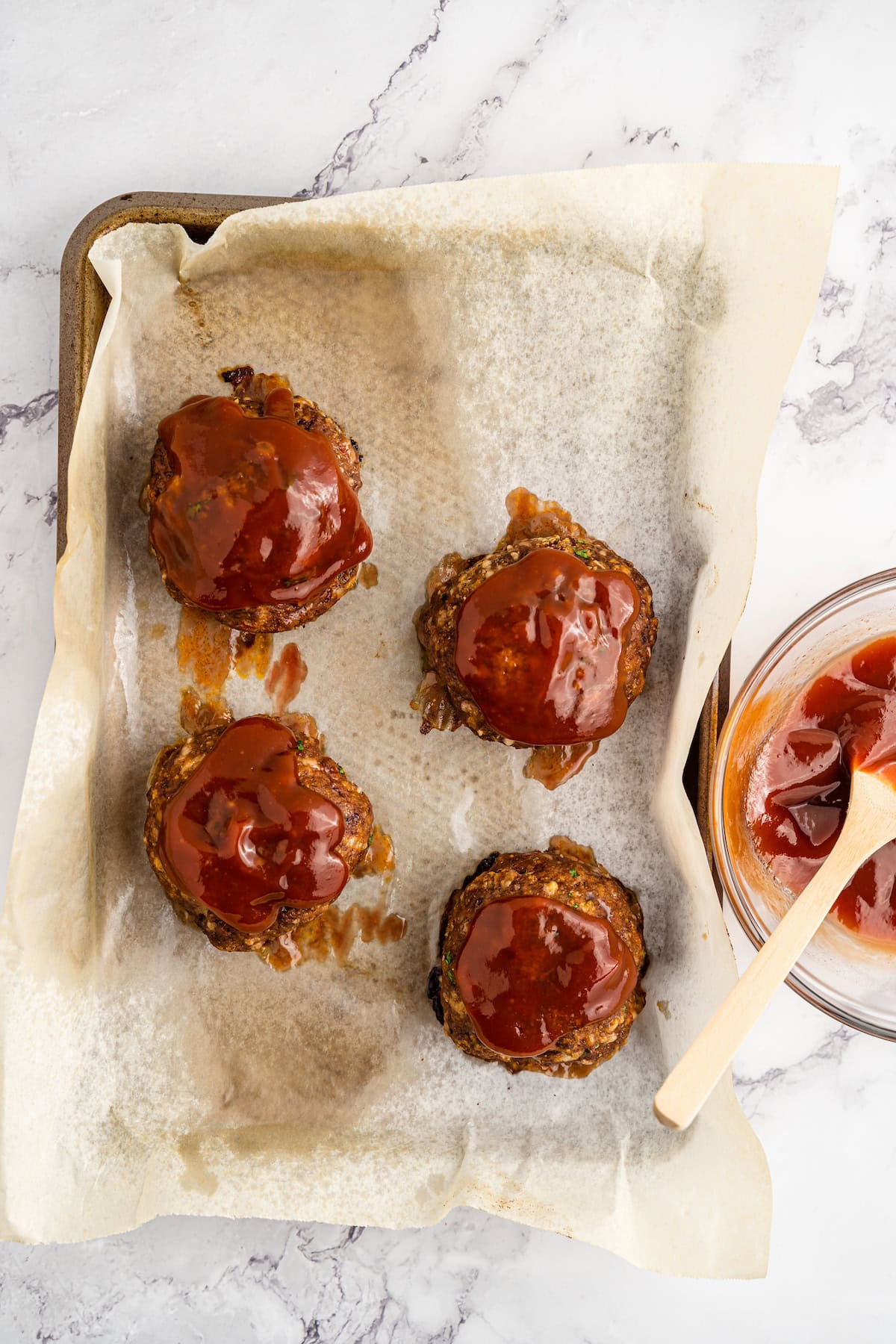 Four mini meatloaves with ketchup glaze on parchment paper, next to a bowl of sauce.