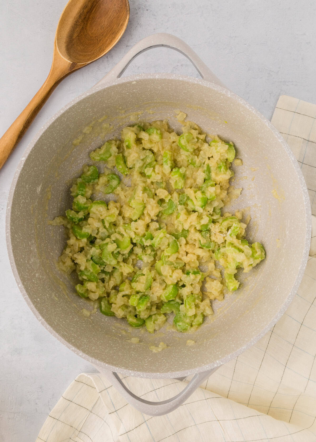 Chopped vegetables sautéing in a light-colored pan next to a wooden spoon and cloth.