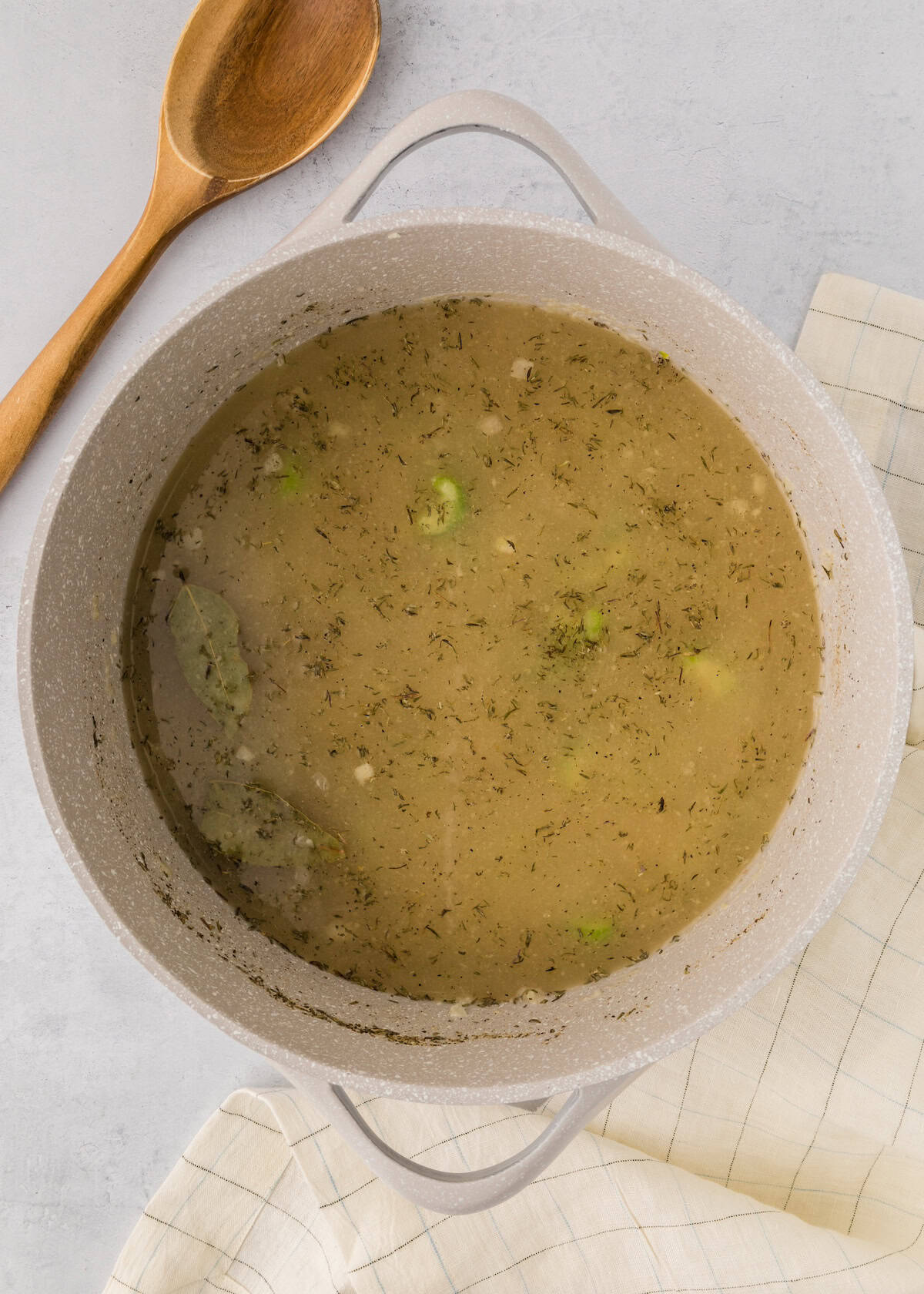 A pot of broth with herbs and vegetables, next to a wooden spoon and a checkered cloth.