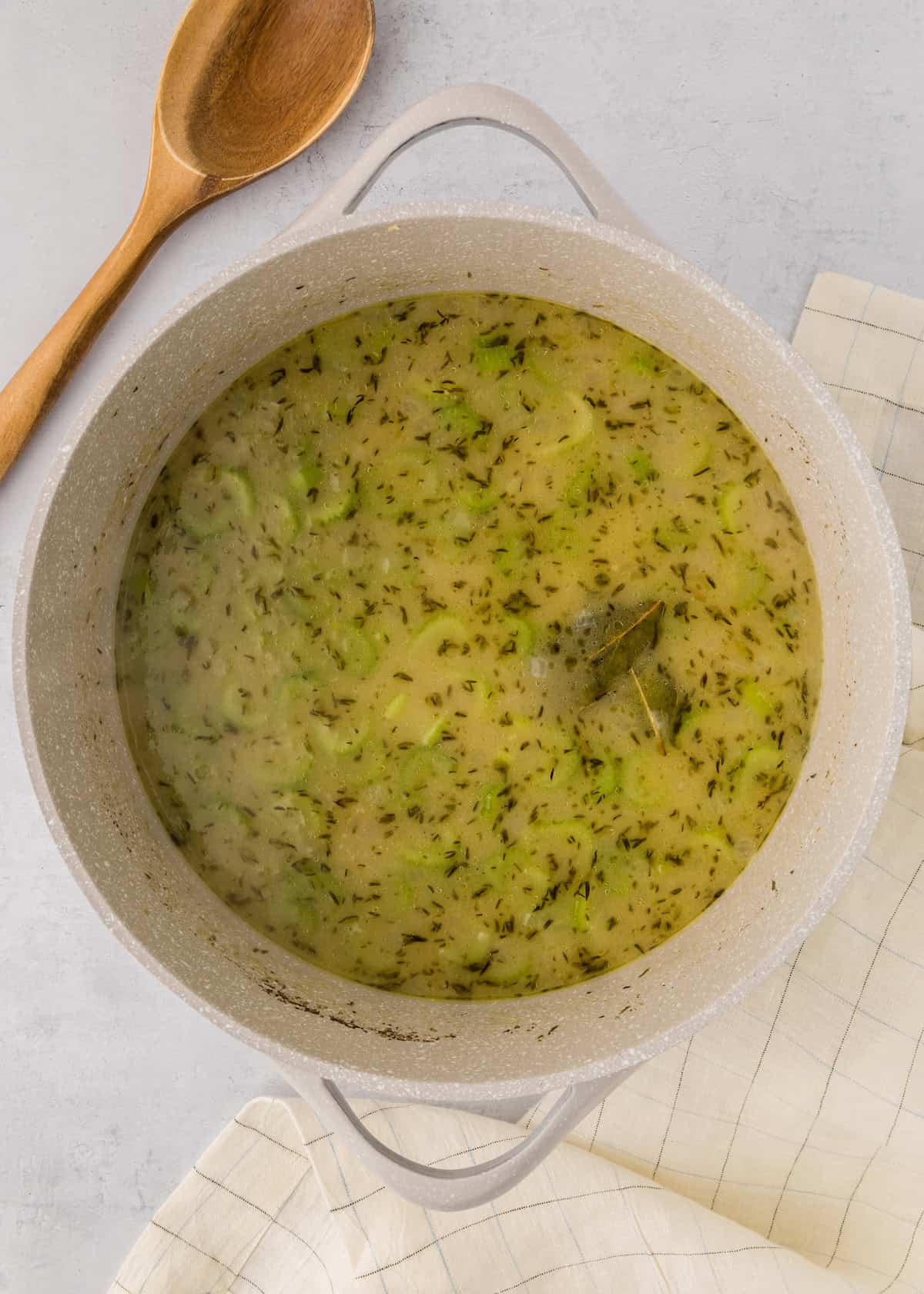 Creamy soup with herbs simmering in a pot, next to a wooden spoon and a white cloth.