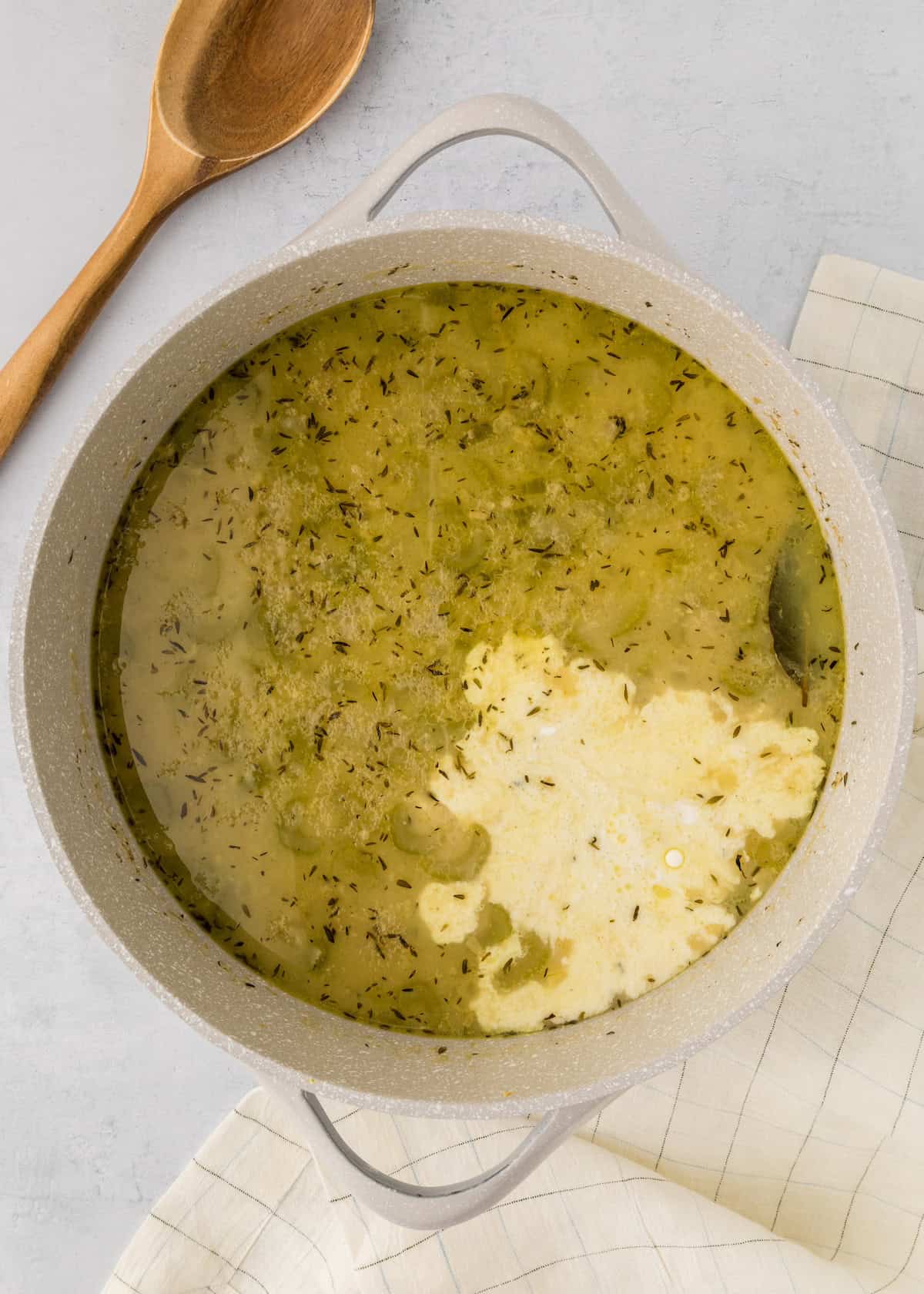 A pot of soup with herbs and a creamy mixture, next to a wooden spoon and cloth napkin.